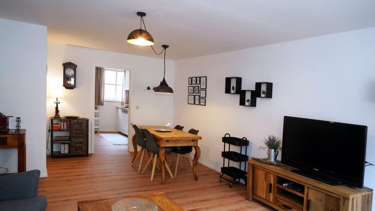 A cozy living room with a dining area and a large television. The bright walls and wooden floor create an inviting atmosphere.