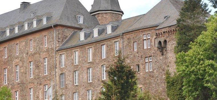 Ein historisches Gebäude mit grauem Dach und Steinfassade. Umgeben von grünen Bäumen und einer malerischen Landschaft.