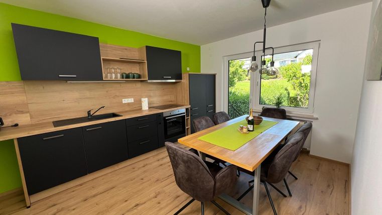A modern kitchen with green walls and black furniture. The dining area is furnished with a large wooden table and cozy chairs.