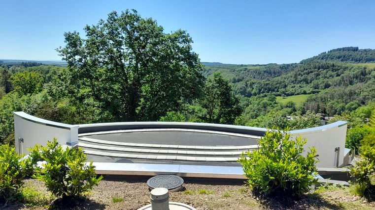 Een modern uitkijkpunt met een halfronde balustrade. Op de achtergrond strekt zich een groen heuvellandschap uit onder een heldere hemel.