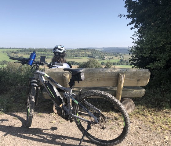 Person mit Helm sitzt auf einer Bank und blickt auf eine weite, grüne Landschaft. Ein Fahrrad steht daneben., © Tourist Information Bitburger Land