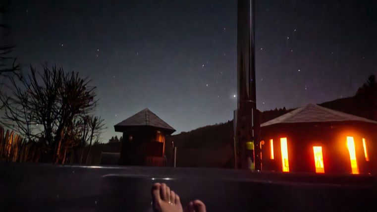 Un bain relaxant en plein air sous un ciel étoilé clair. En arrière-plan, deux bâtiments illuminés et des arbres sont visibles.