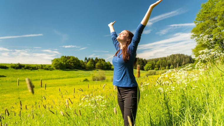 Une femme se tient debout, les bras écartés, dans une prairie verte sous un ciel bleu.