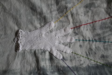 Ein handgefertigter, weißer Handschuh liegt auf einem grauen Hintergrund. Bunte Fäden verlaufen von den Fingerspitzen des Handschuhs in verschiedene Richtungen.