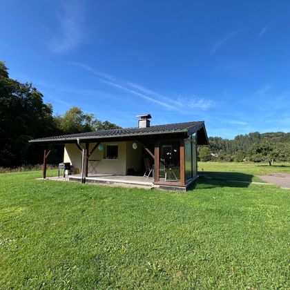 Une petite maison confortable se trouve dans une prairie verte. Le ciel est bleu et clair, entouré d'arbres.