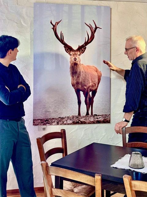 Zwei Männer betrachten ein großes Bild eines majestätischen Rehs mit beeindruckendem Geweih. Das Bild hängt an der Wand in einem gemütlichen Raum mit einem Tisch und Stühlen.