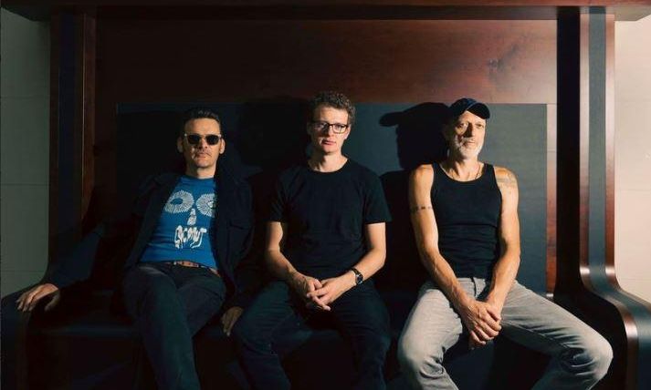 Three men are sitting on a bench. They are wearing casual clothes and looking at the camera.