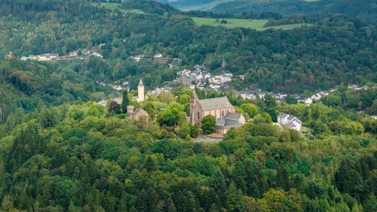 A picturesque landscape with an old church, surrounded by dense forests. In the background, gentle hills and a small village can be seen.