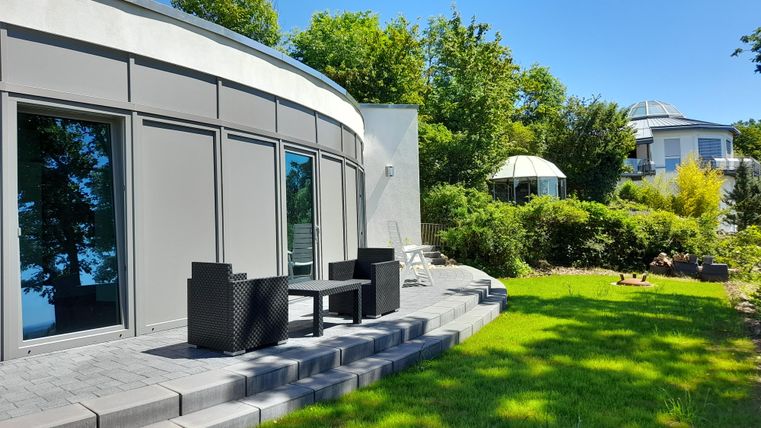 Een moderne woning met grote ramen en een goed onderhouden tuin. Op het terras staan zwarte tuinmeubelen en het is zonnig.