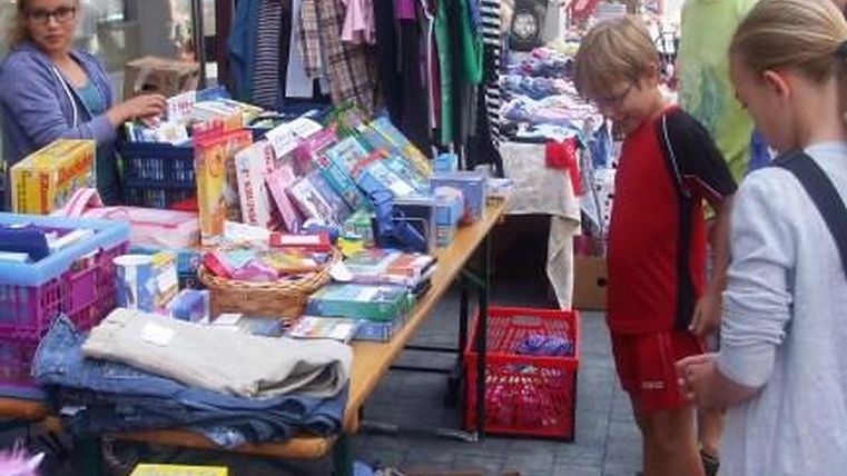 Bunter Flohmarktstand mit Kleidung, Büchern und Spielzeug. Einige Personen stehen davor und betrachten die Ware.