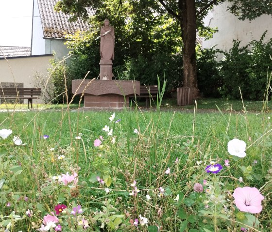 Eine Statue von Donatus steht in einem Garten. Im Vordergrund blühen bunte Blumen. Bänke und ein Baum sind im Hintergrund zu sehen., © TI Bitburger Land