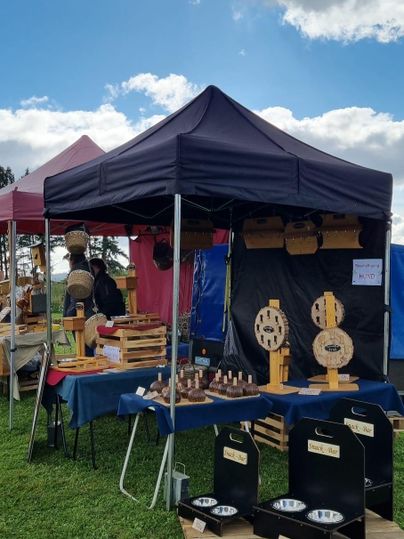 Ein Marktstand mit einem schwarzen Zelt und handgefertigten Holzprodukten. Im Hintergrund sind weitere Stände und ein blauer Himmel mit Wolken zu sehen.
