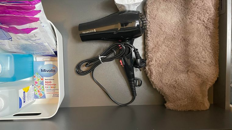 Un sèche-cheveux est posé sur un support avec une serviette et divers produits de soin. À côté se trouve un bol blanc et un tissu doux et brun.