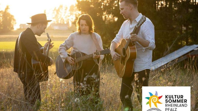 Drie muzikanten spelen harmonieus gitaar op een veld bij zonsondergang. De sfeer is ontspannen en creatief.