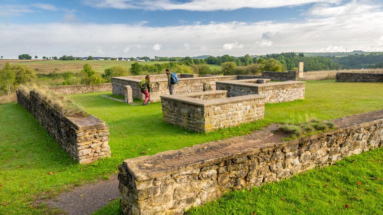 Ruinen einer alten Anlage auf einer grünen Wiese mit Blick auf die Umgebung. Zwei Personen erkunden die historischen Steine.