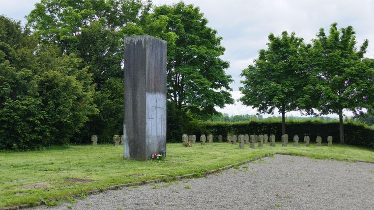 Op een groene weide staat een stenen monument, omringd door bomen. Rondom het monument liggen kleine steentjes.