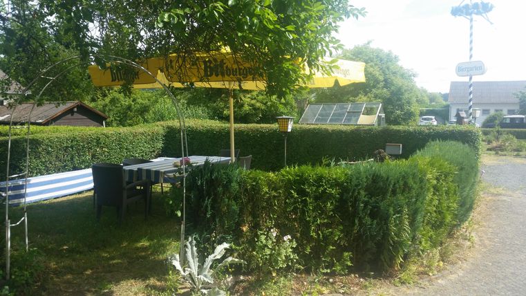 Ein einladender Außenbereich mit einem Tisch und Stühlen unter einem gelben Sonnenschirm. Grüne Hecken und Pflanzen umgeben die gemütliche Sitzecke.