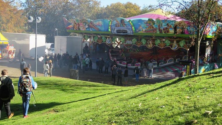 A fair with rides and many people. In the background, colorful stalls and autumnal trees can be seen.