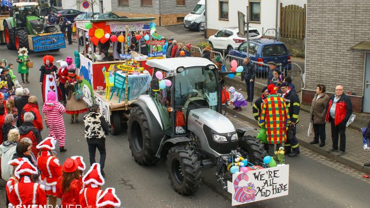A colorful parade with a tractor and festively decorated floats. Clowns and people in red costumes are joyfully celebrating on the street.