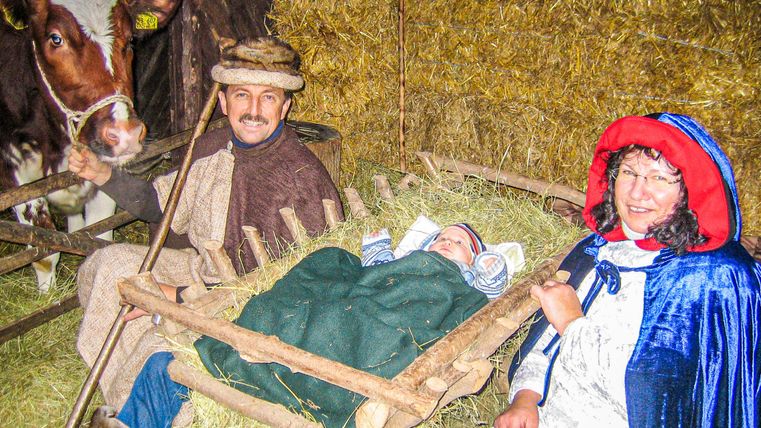 Eine Familie sitzt in einem Stall mit Heu und einer Kuh. Ein Baby liegt in einer Wiege, während die Eltern daneben lächeln.