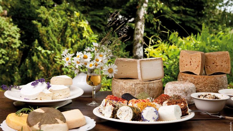 Een selectie van verschillende soorten geitenkaas op een tafel in de buitenlucht. Op de achtergrond versieren bloemen en groen loof de scène.