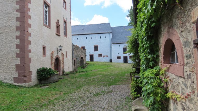 Eine malerische Ansicht eines Innenhofs mit historischen Gebäuden und üppiger Vegetation. Der Hof ist mit Kopfsteinpflaster ausgelegt und von alten Mauern und weißen Gebäuden gesäumt, die mit Efeu bedeckt sind.