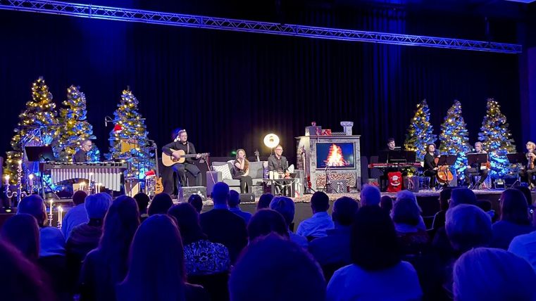 Une représentation scénique festive avec des musiciens et des arbres décorés pour Noël. Le public est attentif au premier plan.