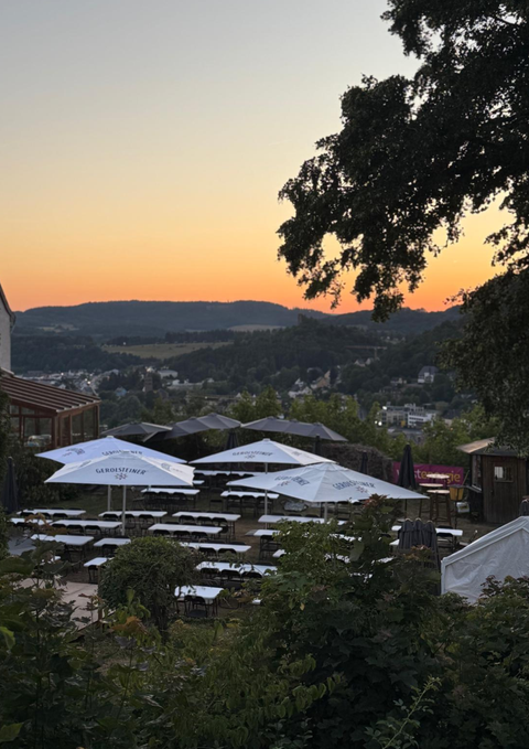 Am Abend sitzen leere Tische und Stühle unter Sonnenschirmen in einem Biergarten. Im Hintergrund ist eine hügelige Landschaft mit einer Stadt zu sehen, die im warmen Licht des Sonnenuntergangs erstrahlt.