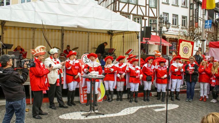 A group of people in red uniforms is standing on a stage and singing. In the background, historical buildings and a tent roof can be seen.