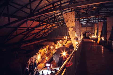 Eine moderne Ausstellungshalle mit einem Holzsteg und sanfter Beleuchtung. Im Hintergrund sind archäologische Funde und Besucher zu sehen.