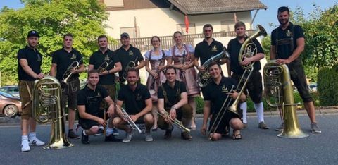 Eine Gruppe von Musikern steht in einer Straße. Sie tragen Trachten und halten verschiedene Blasinstrumente in den Händen.