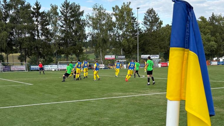 A soccer game on a green field with players in yellow-blue jerseys. In the foreground, a flag in blue and yellow flutters.