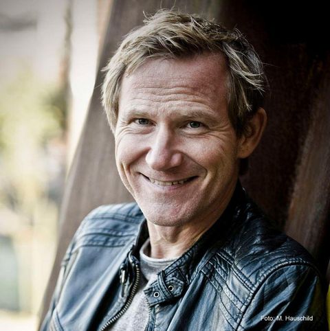 A smiling man in a leather jacket in front of a blurred background landscape. He has short, blonde hair and a friendly face.