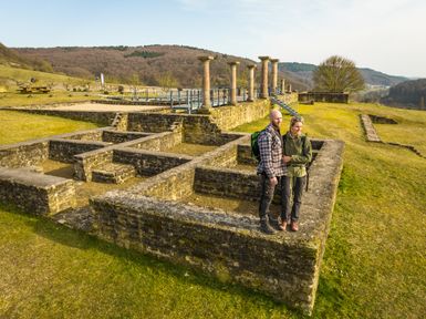 Zwei Besucherinnen erkunden die freigelegten Grundmauern der römischen Villa Holsthum mit sichtbaren Steinmauern und Säulenresten, eingebettet in die hügelige Kulturlandschaft der Südeifel bei sonnigem Wetter.