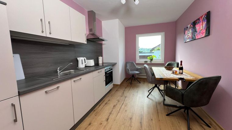 A modern kitchen with bright furniture and a dining area. The walls are in soft shades, and a window lets in plenty of daylight.