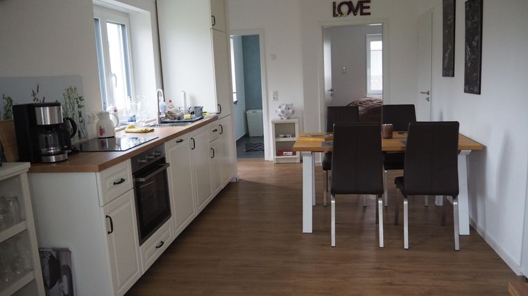 Eine helle Küche mit modernen Möbeln und einem Essbereich. Der Raum hat Holzfußboden und große Fenster für viel natürlichen Licht.