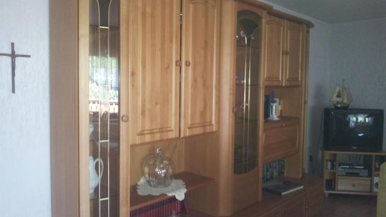 Ein Holzschrank mit Glastüren und Dekorationsobjekten. Im Hintergrund ist ein Fernseher und eine Ecke des Raums sichtbar.