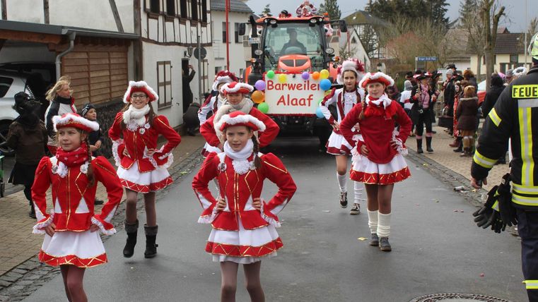A joyful carnival parade with costumed dancers in red outfits. In the background, a festively decorated float can be seen.