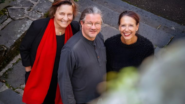 Drei Personen stehen lächelnd auf einer Treppe. Sie tragen verschiedene Kleidungsstile und wirken freundlich und entspannt.