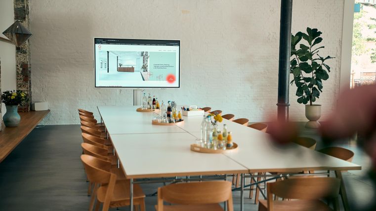 A modern conference room with a long table and wooden chairs. A screen displaying presentation content is mounted on the wall.
