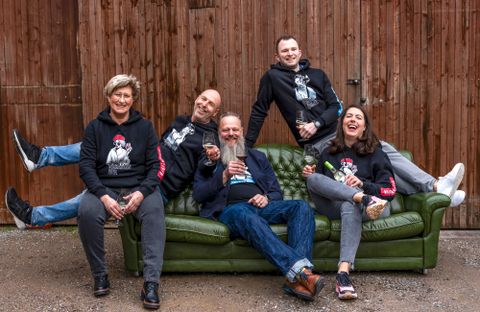Eine Gruppe von fünf Personen sitzt und lehnt sich entspannt auf einer grünen Couch. Sie lachen und halten Getränke in der Hand, während sie lässige Hoodies tragen.
