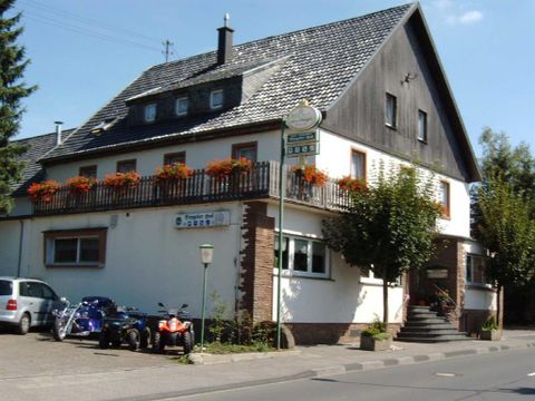 <p>Een drie verdiepingen tellend gebouw met een zwart dak. De ingang is te bereiken via een trap. Op de eerste verdieping omringt een houten leuning met bloembakken een groot balkon. Links naast het huis is een parkeerplaats, die bezet is door drie auto's. Achter de parkeerplaats hangt een bord met de tekst "Biergarten". Voor het hotel loopt een stoep die direct aan de hoofdweg grenst.</p>