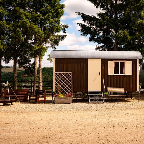 Une petite maison en bois se trouve sur un terrain de gravier entouré de grands sapins. Il y a quelques places assises et une ambiance détendue.