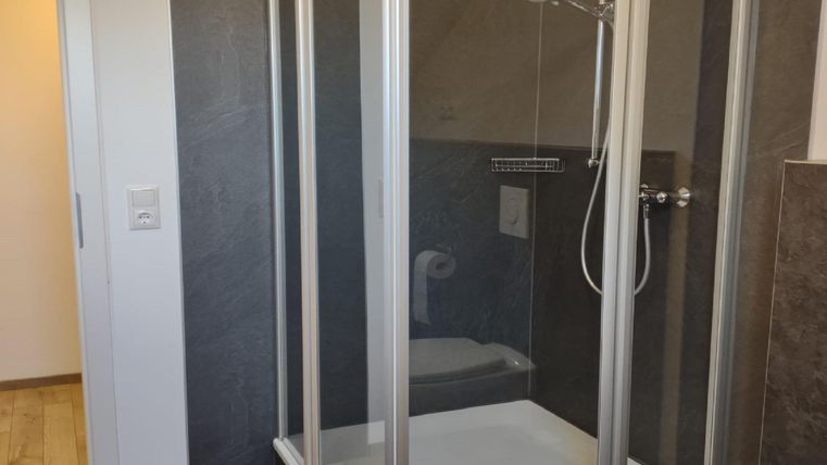 A modern shower cabin with clear glass walls is in a bright bathroom. The floor is tiled with wooden planks, and there is a door leading to another room.