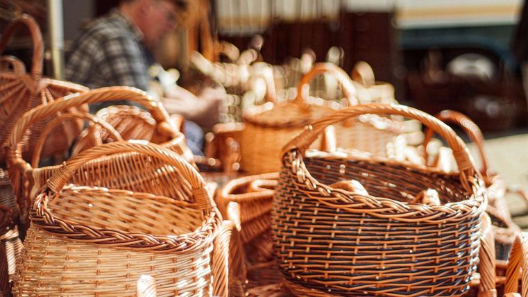 Handgemaakte rieten manden in verschillende maten staan op een tafel. Op de achtergrond werkt een ambachtsman aan andere manden.