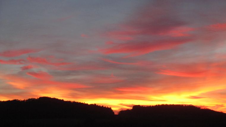Een adembenemende zonsondergang met felrode en oranje wolken. Op de voorgrond zijn de silhouetten van heuvels te zien.