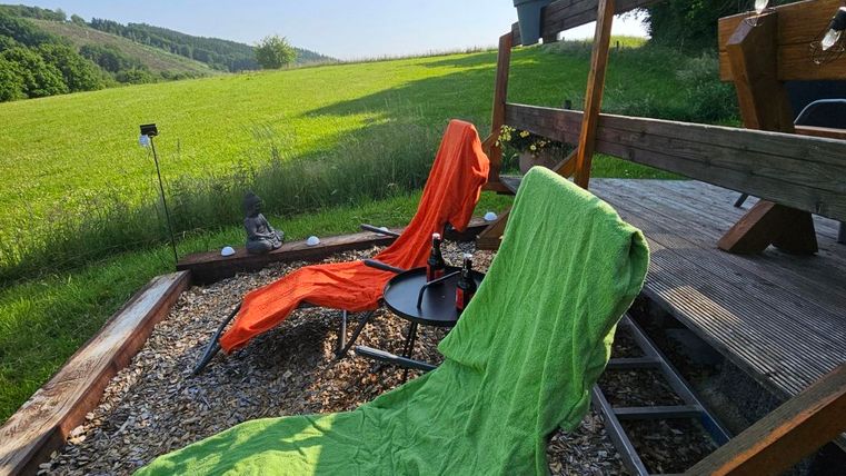 Two lounge chairs in bright colors are standing on a wooden deck. In the background, a green meadow stretches under a clear sky.
