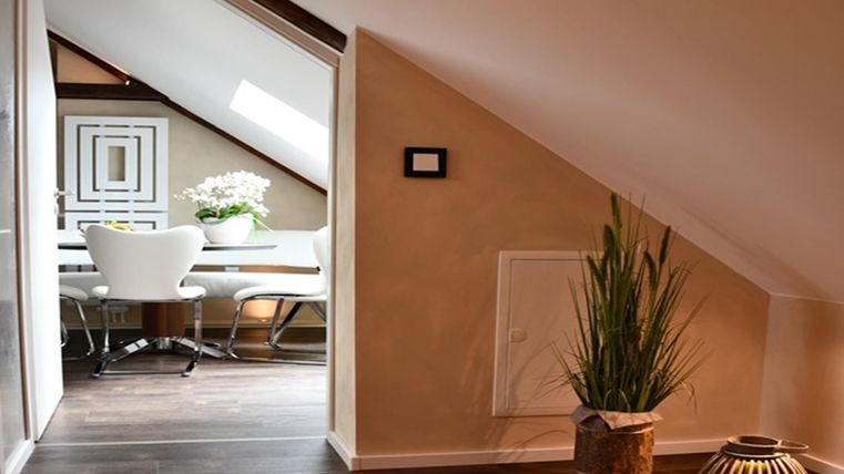 A cozy attic space with slanted walls. In the background, a dining area with a table and chairs can be seen.