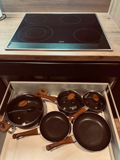 Une armoire de cuisine avec plusieurs poêles et casseroles, qui ont des poignées en bois. En arrière-plan, il y a une plaque de cuisson en vitrocéramique.