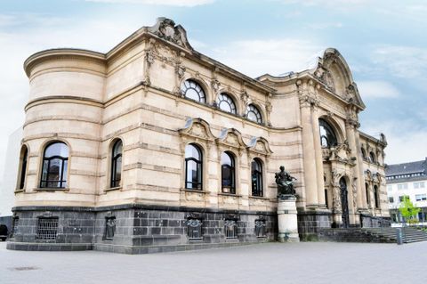 Ein historisches Gebäude mit einer beeindruckenden Fassade und großen Fenstern. Im Vordergrund steht eine Statue, die dem Gebäude einen besonderen Akzent verleiht.
